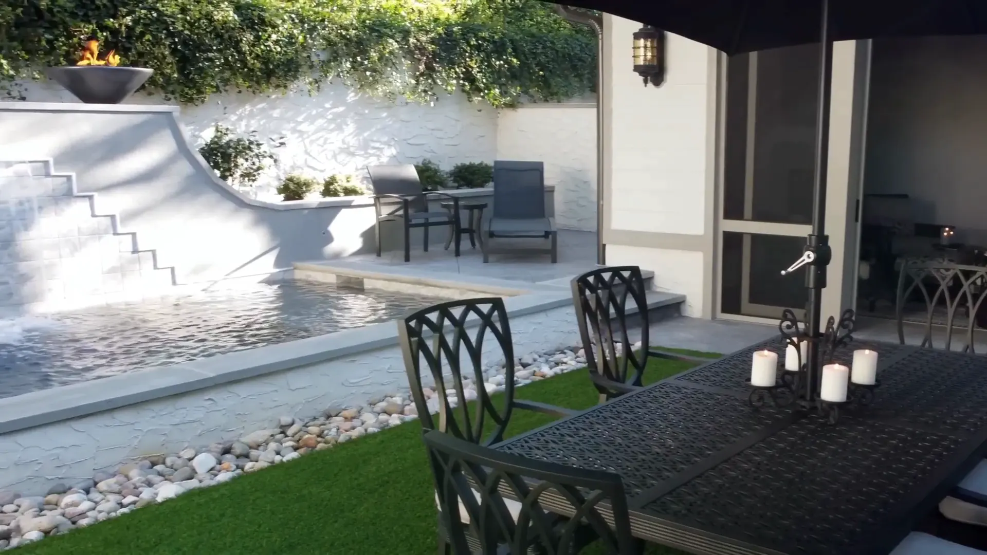A covered dining patio with a long table beside a pool and a tiled water wall